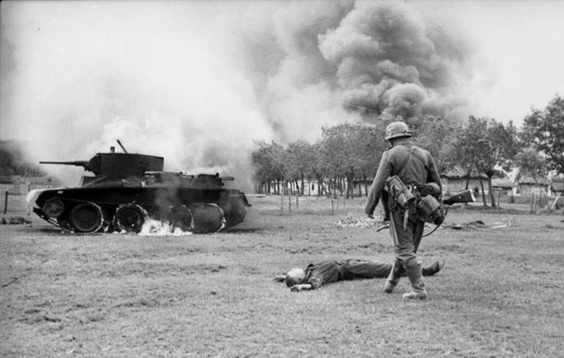 Немецкий солдат и лежащий на земле труп советского солдата у горящего танка БТ-7. 1941г