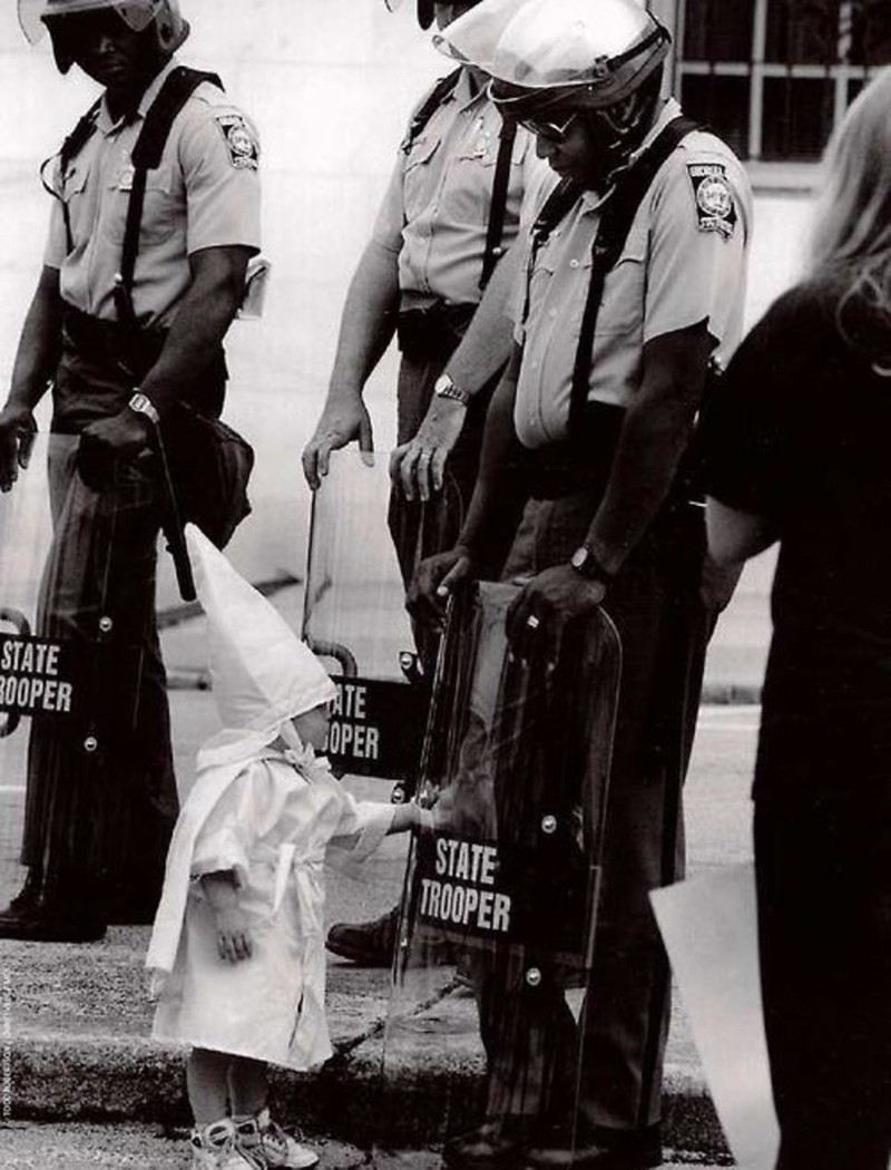 Ребенок члена организации Ку-клукс-клан во время демонстрации дотрагивается до своего отражения в полицейском щите, который держит полицейский-афроамериканец (1992 г.)
