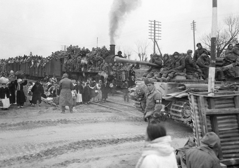 Поезд с беженцами стоит у переезда, пропуская британский бронетранспортёр, недалеко от Осана, Южная Корея, 15 января 1951 года. 