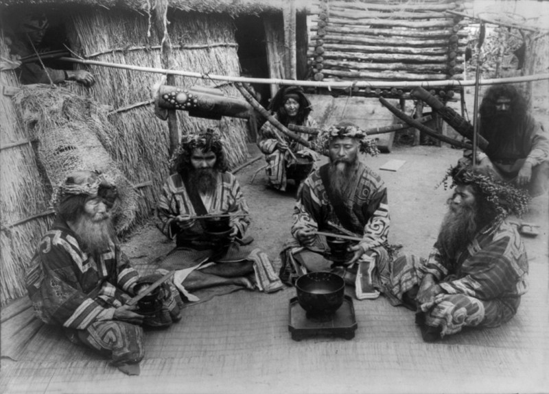 Шесть айнов в традиционных нарядах сидят у чаши. 