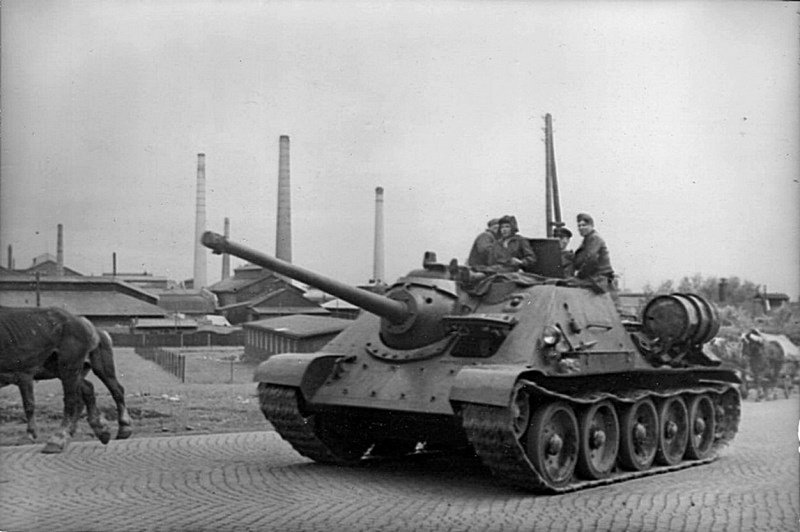 Советские солдаты на броне САУ СУ-85 проезжают по дороге немецкого населенного пункта.