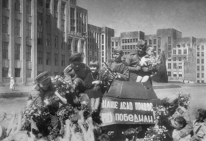 Маленькие жители Минска поздравляют советских бойцов с Победой у бронеавтомобиля БА-64 на площади Ленина со зданием Дома правительства.
