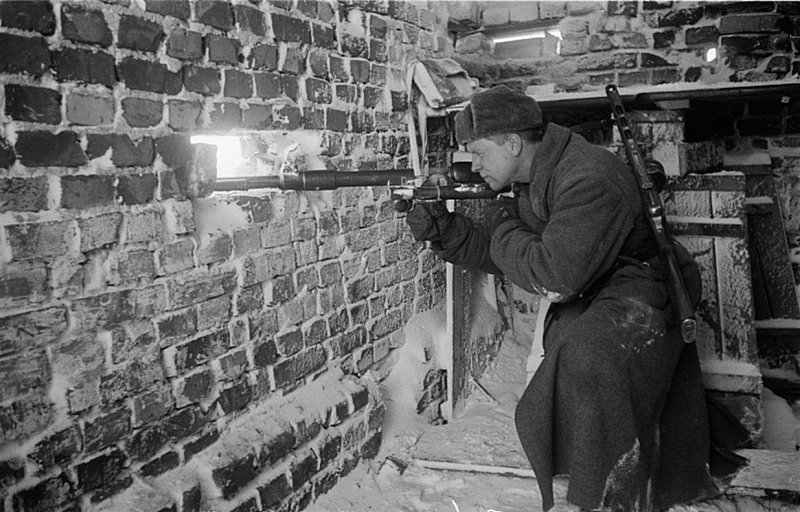 Советский бронебойщик, вооруженный 14,5-мм противотанковым ружьем системы Дегтярева, образца 1941-го года (ПТРД-41) прицеливается через стену здания (за плечами пистолет-пулемет ППШ). На полке у стены расположена брезентовая патронная сумка на 20 пат