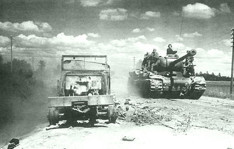 Танк ИС-2 с десантом на борту движется по Рижскому шоссе. На обочине дороги разбитый и сгоревший грузовик Studebaker US6. сентябрь-октябрь 1944 