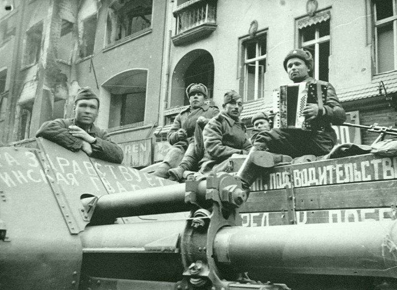 Советские артиллеристы слушают игру на аккордеоне своего товарища на улице Берлина. На переднем плане — 122-мм гаубица М-30.