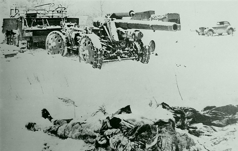 Тело убитого немецкого солдата в снегу у 150-мм тяжелой полевой гаубицы (15 cm sFH 18). За гаубицей находится немецкий разукомплектованный полугусеничный тягач с номером подразделения войск СС. Фотография сделана в период битвы за Москву.