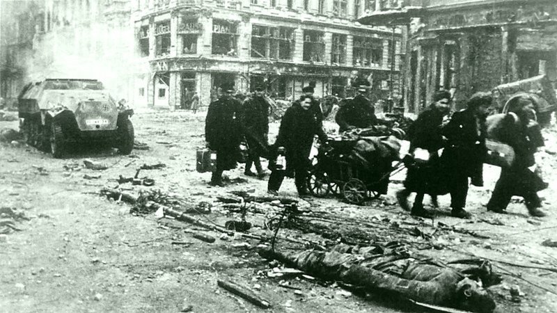 37. Немецкие беженцы, среди которых находятся мужчины в форме, проходят рядом с телом убитого солдата немецкой армии на улице Берлина.