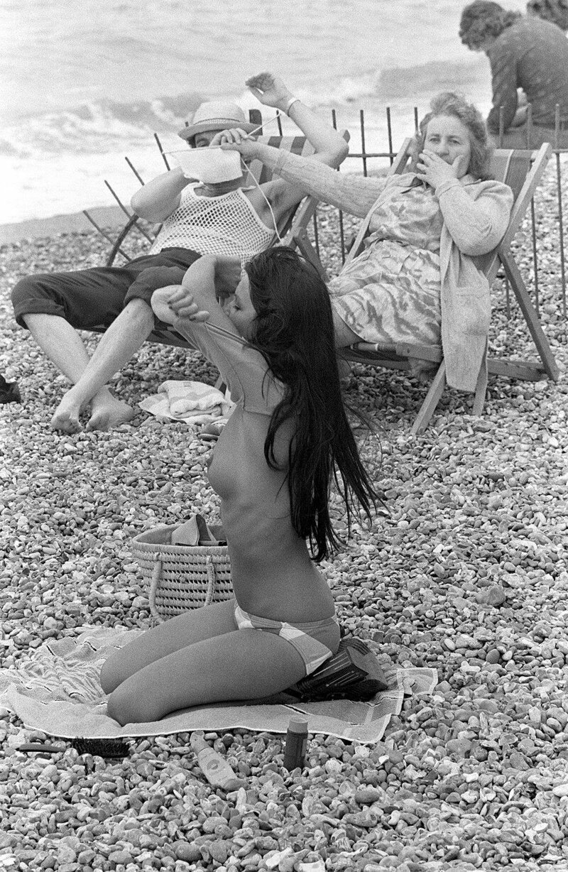 Женщина загораживает обзор мужу, чтобы тот не смотрел на раздевающуюся на пляже девицу. 