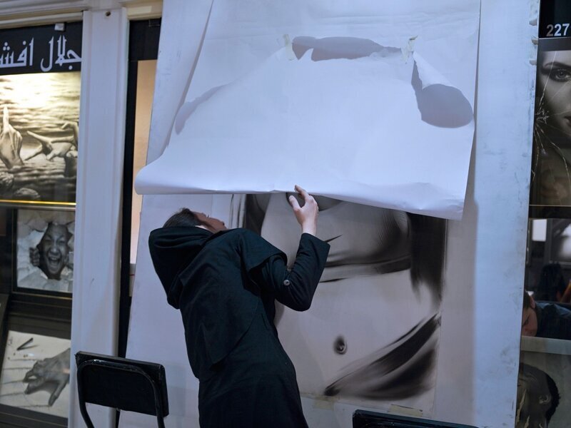 Девушка пытается посмотреть на фотографию женщины, скрытую под белой бумагой. По исламским законам Ирана запрещено показывать женские тела на публике.