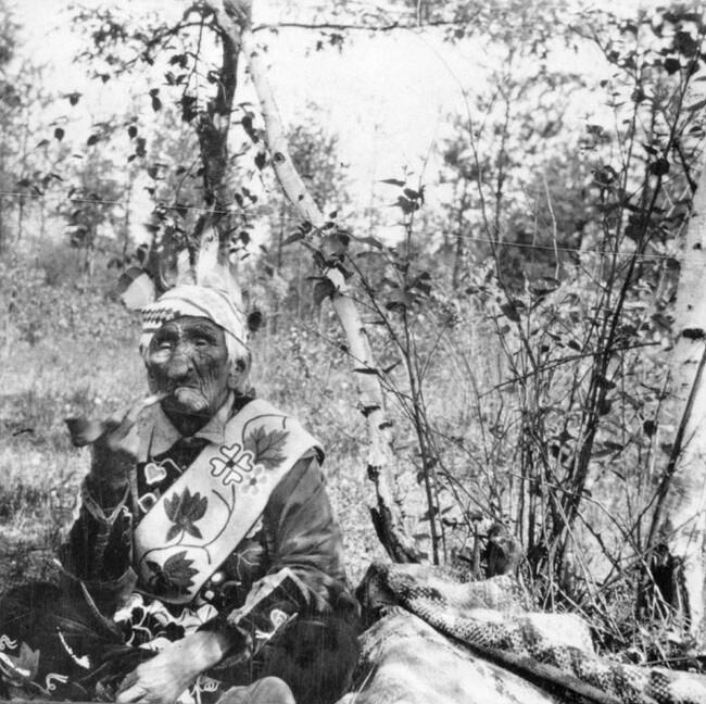 Белый Волк курит трубку, сидя в лесу в традиционной индейской одежде, примерно 1910-е годы