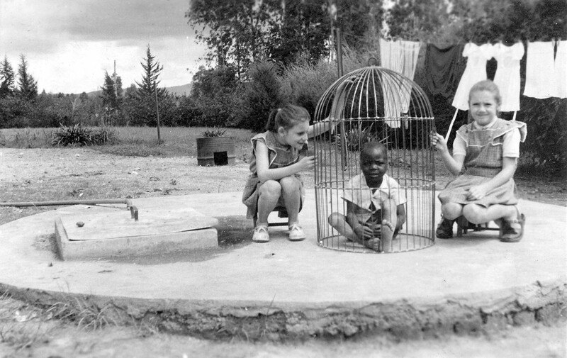 Две белые девочки играют с негритёнком в клетке для птиц, Бельгийское Конго