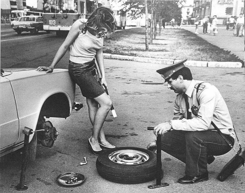 Сотрудник ГАИ помогает девушке заменить колесо пока она держит его жезл. СССР, 1981 год