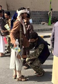 Что значит, что он пытается найти? Оружие разумеется. А автомат на плече - самого охранника/полицейского. Он просто попросил его подержать, пока руки обыском заняты. Разве не понятно?