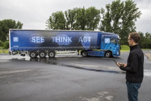 Это не ново... 
На картинке, как вы скажете - фура. Для облегчения маневрирования в стесненных условиях (например, грузовой терминал), водитель может выйти из кабины...
Устанавливаемое на планшет (обычный планшет!) приложение позволяет задавать скорость при движении вперед (три режима) в 4, 2 или 1 км/ч, при движении назад (еще три режима) - 2, 1 или 0,5 км/ч. При этом грузовик движется, используя только электропривод, с минимальным уровнем шума и нулевым уровнем вредных выбросов. Канал беспроводной связи использует технологию Bluetooth-Low-Energy (BLE) с дальностью действия до 25 метров. Теоретически запаса хода на аккумуляторах хватает на 10 полных маневров, связанных с подъездом к погрузо-разгрузочной площадке терминала (разумеется, если грузовик не был припаркован в соседнем квартале). BLE поддерживается смартфонами, начиная с iPhone 4S и планшетами, начиная с iPad 3, равно и альтернативными решениями от Google, начиная с использующих версию Android 4.3 и Microsoft (Windows Phone 8.1). Кроме того на машине установлено 6  (полуприцеп) или 8 (прицеп) видеокамер. 
Управление с планшета очень простое. На экране схема грузовика, совмещенная с "картинками" бортовых видеокамер. Приложили палец к экранной кнопке ("D", "N", "R") - машина начала движение вперед или назад, потянули в сторону (экранные кнопки-стрелки) - начинается поворот. Сняли палец - машина останавливается. Упомянутые видеокамеры посильно контролируют ситуацию вокруг грузовика (хотя картинка мелковата), так что "бегать кругами" вокруг машины водителю не нужно. По соображениям безопасности при потере связи грузовик мгновенно останавливается.
Теперь вес. Все вместе: гибридный привод, преобразователь, накопители увеличивают снаряженную массу на 350 кг. Эту цифру надо минусовать из цифры коммерческой загрузки. Но для 40 тонн это не всегда критично - легковесные объемные грузы на таких машинах преобладают, будет чугун: да, обидно, аж на две-три чушки меньше...
Разумеется это опция и на прототипе цена порядка 2500 евро (сам автопоезд с картинки порядка 150-160 тысяч). Все будет доступно для заказа начиная с 2018 года и в серии возможно цена снизится. Хотя на самом деле и нынешняя не очень высокая. Ибо если ваш наемный водитель на терминале зацепит чужую машину, то ремонт двух машин может быть за ваш счет. Страховка не имеет значения: когда виновник понятен, будет регресс. Да и в следующий раз ставка вырастет. Кроме того есть понятие упущенной выгоды (машина не вела груз, а стояла в ремонте), да и грузовладелец, особенно если груз срочный, тоже может выкатить сумму претензий, многократно превышающую стоимость опции... Требование компенсации убытков по последней причине может предъявить и владелец машины, которую задел ваш водитель. Так что считайте сами...