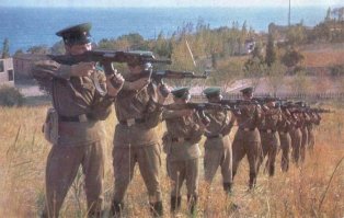 при стрельбе стоя (особенно в движении) для большей устойчивости и надёжности хвата левая рука прижата к корпусу, по этому и автомат удерживается за магазин, а не за цевьё. вполне нормальное явление в практической (не спортивной) стрельбе.