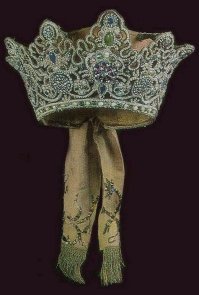 Коруна. Вырезалась из той же бересты что и венец, но с с зубцами по краю. Украшалась полудрагоценными и даже драгоценными камнями, жемчугом, бисером, перламутром. Надевалась на свадьбу. Кто не мог себе позволить- брал в аренду, могла быть одна коруна на всю волость. Те, кто побогаче передавали от матери к дочери на протяжении многих поколений в качестве приданного.
Иногда коруны были очень высокие, а с учетом деревянного основания- тяжелые, на голове держались чисто символически. Поэтому были даже придуманы специальные бабы, которые с невестой рядом ходили и всю эту красоту придерживали.