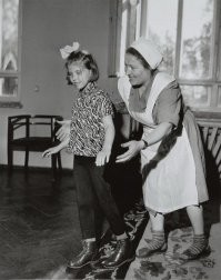 Некорректное название. Это не воспитательница, а нянька.

Обратите внимание на обувь девочки: это специальные ортопедические ботинки. Они помогали в реабилитации некоторых жестких проблем с опорно-двигательного аппарата (мне такие пришлось год таскать после сложного перелома голеностопа в 4 года) и облегчали возможность ходить детям с ДЦП (до 10...12 лет).