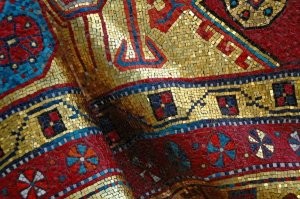 Может показаться, что могила знаменитого танцора Рудольфа Нуриева во Франции укрыта настоящим старинным ковром. Многие туристы даже спрашивают – не вредит ли ковру дождь… Дождь ему не страшен – ведь надгробие сделано из камня и украшено тончайшей мозаикой.
В 1996 году надгробие было изготовлено в итальянской мозаичной мастерской Акомена Спацио Мозаико (Akomena Spacio Mosaico). Мозаика ковра выполнена из мелких преимущественно квадратной формы элементов с плотнейшим прилеганием деталей, практически без видимых швов. Но при этом поверхность мозаики оставлена шероховатой, с весьма резкими изменениями уровня мозаичных элементов. Этот прием с расстояния уже 2-3 метров создает общее впечатление ковровой текстуры.