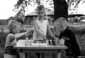 Странное фото. 
Игра вроде серьёзная, судя по часам контроль не менее часа на партию, а за доской пацаны до 10 лет.
Игра ещё не вышла из дебюта, а какой-то тип уже лезет с подсказками.
Ну и главная несуразность, играют пацаны в шахматы, а коробки рядом с ними стоят с надписью "Шашки". Причём сразу два комплекта, коих явно много для одной доски.