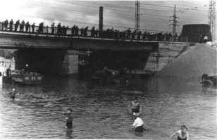Снимок №5 "Потоп на пл.Труда" из серии снимков 1959 года сделаных жителем Восточного посёлка (это адрес такой) Георгием Викторовичем Жаннин-Перро. Он вообще много снимал Новосибирск и вот часть этой серии. В тот летний день с юго-востока пришла чёрная, налившаяся над Обью, избытком воды, она сначала обрушила водяной шквал на строящийся район "Башня", а потом накрыла Первый соцгород, где все переулки и улицы были мощёны булыжником, а центральную улицу Станиславского упиравшуюся в только, что построенный и открытый новый путепровод под Транссибом, свеже заасфальтированную для пуска троллейбусного движения к заводам.  Ливнёвка на площади Труда в мгновение ока была забита обломками смытого асфальта и площадь превратилась в озеро. Но кому проблемы, а детям и тут игра - вода-то пришла тёплая, нагретая взломанным дорожным покрытием. И вот благодаря Георгию Викторовичу, мы можем увидеть результат работы того ливня.