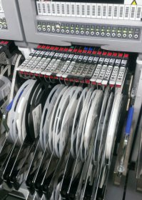 Там фотография всего цеха была и если эта тётушка паяет образец, то кроме образцов тот цех ничего не выпускает. Отличие фоток с Gigabyte достаточно очевидно - во-первых там конвейер. Во-вторых те, кто занят ручным трудом, расставляют на плату крупные компоненты, поставляемые  россыпью. С SMD к этому моменту уже закончили, а выглядело это как на картинке ниже (картинка с того же завода). Катушки, подвешенные снаружи автоматов, как раз содержат ленты с SMD.
Причём Gigabyte это ещё не самое технологичное производство - там, например, контроль осуществляется визуально, в то время как на большинстве других заводов изображение платы с установленными компонентами анализирует компьютер.