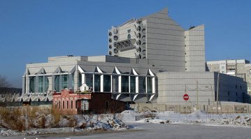 «Бастилия» — культовое здание Благовещенска, здание-монстр. Одной из последних ее жертв был маленький кирпичный домик. Вспоминая о его злосчастной судьбе, трудно поверить в то, что «Бастилия» закрыла этот свой черный список.

Последняя жертва «Бастилии»

Общественно-культурный центр не имеет официального названия даже спустя два года после открытия. Назвать его «Бастилией» власти не могли, а другого названия сама «Бастилия» не принимает, это имя она выбрала себе сама и так ее зовет народ. А ведь сколько конкурсов проводилось по этому поводу, сколько комиссий совещалось…

Для тех кто не в теме кратко скажем, что это здание некогда было самым известным долгостроем Благовещенска. Его железобетонные конструкции десятилетиями возвышались в центре города. В лунную ночь «Бастилия» выглядела не просто зловеще, она внушала ужас.

Её начали строить глубоко в СССР, еще до перестроек. Тогда она была «драматическим театром». Ее строили так медленно, с такими проволочками, что даже проплывавший на пароходе по Амуру какой-то советский писатель, живописуя рассвет социализма, написал, как огнями ослепило его здание драмтеатра на берегу реки. И хотя он видел лишь остов здания, его книга была издана много позже путешествия, и автор не мог представить себе, что здание до сих пор не достроено. Вышел конфуз.

Заброшенная стройка стала местом прибежища бомжей, местом тусовки неформальной молодежи, притоном наркоманов. Там то и дело находили трупы, упавших с конструкций, убитых кем-то или просто умерших. Периодические трагедии в «Бастилии» поднимали мелкие волны общественного негодования, но ничего не менялось, строительная площадка зарастала не просто бурьяном, а диким лесом уже.

Иногда возникали проекты то по ее достройке, то по сносу. Но все было впустую, менялись времена, люди, нравы, а «Бастилия» оставалась сама собой. Сколько уж там разных жертв на ее счету, поломанных судеб, то уж неизвестно и не сосчитать. Бывший губернатор Коротков рискнул ее достроить, и с перерывами стройка велась, но и ему не суждено было изменить ее суть. Скорее наоборот: перед самым открытием Короткова сняли, а утвержденное им название ОКЦ — «Албазин» следующий губернатор отменил и другого уже не давали.

Похоже, попытка обозвать «Бастилию» сыграла мистическую роковую роль в судьбе самого Короткова: утвердил название и через три дня получил указ Президента об отстранении. Не любит «Бастилия» такого вот панибратства. Может на ее месте было какое-нибудь священное место древних джурдженей? Очень на то похоже.

Конечно, она давно расчистила пространство вокруг себя, целый городской квартал снесли в свое время ради нее, но каким-то чудом совсем рядом сохранился маленький, еще дореволюционный домик. Почему его не снесли в советские времена, как он остался стоять рядом с монстром все эти десятилетия – известно теперь только самой «Бастилии». И уже одевшись в белый мраморный саван, она словно оглядевшись и увидев у себя под ногами это несчастный домик – как всегда жестко и безжалостно, одним ударом прикончила и его.

Формально дело было так.

Власть в области сменилась, на место Короткова, был назначен Колесов, а тут как раз и подоспело официальное открытие ОКЦ. Новый губернатор поехал посмотреть «объект» и увидел красный домик, явно мешавшийся под ногами у «Бастилии» и портивший вид. Приказал немедленно снести.

Дело в том, что избавиться от домика пытались и до Колесова, но он находился в частной собственности, им владела некая гражданка по фамилии Левцкая, которая толи хотела открыть там кафе, толи просто пыталась получить побольше денег с города, раз уж такая сложилась ситуация. В общем, шли затяжные переговоры с собственником домика о цене.

Возможно, они бы шли и до сих пор, если бы не назначение Колесова, который отличался своеобразным взглядом на мир. В общем, он дал мэру Мигуле неделю на решение вопроса и уехал. А Колесова тогда все сильно испугались, противоречить ему не смели, он был страшнее любого закона для посткоротковской чиновной братии.

Сына той самой гражданки Левицкой вызвали к вице-мэру, который предложил ей домик этот быстро подарить городу, мол, а то хуже будет. Гражданка отказалась. Ночью к домику с милицейским сопровождением подъехал «Камацу», собственница с родственниками собирались держать оборону, но их быстро под белые рученьки спровадили и экскаватор раскрошил домик вместе со всем содержимым. К утру все было кончено.

Теперь лишь фотографии снятые в свое время со стороны Амура напоминают об этом злосчастном доме, а «Бастилия» стоит на берегу Амура в гордом одиночестве.

Мы не будем судить о соблюдении в нашем городе законности, о правомерности и адекватности действий различных людей. В этой рубрике мы рассказываем о Благовещенске и его городских легендах. «Бастилия» — одна из них. Не по своим масштабам, ни по архитектуре она никогда не соответствовала тихой провинциальной столице Приамурья. История ее появления загадочна, а ее судьба по-своему уникальна.

Находись такое здание в каком-нибудь туристическом центре, оно бы стало местом паломничества любопытных. «Бастилию» недаром так назвал народ, она коверкает судьбы связавшихся с ней людей не хуже своей французской тезки. Только ту разрушали, но в своей новой реинкарнации на берегу Амура символ зла продолжает жить.

Маленький красный домик, стал очередной жертвой «Бастилии», но никто не поверит, что последней. Сейчас там выступают заезжие «биланы» и все кажется вполне благополучным. Но если завтра рухнут, признанные абсолютно надежными после двадцати лет стояния под снегом и дождем, ее перекрытия, или вспыхнет пожар, или случится что-нибудь не менее ужасное, то разве кто-нибудь в Благовещенске этому удивится?